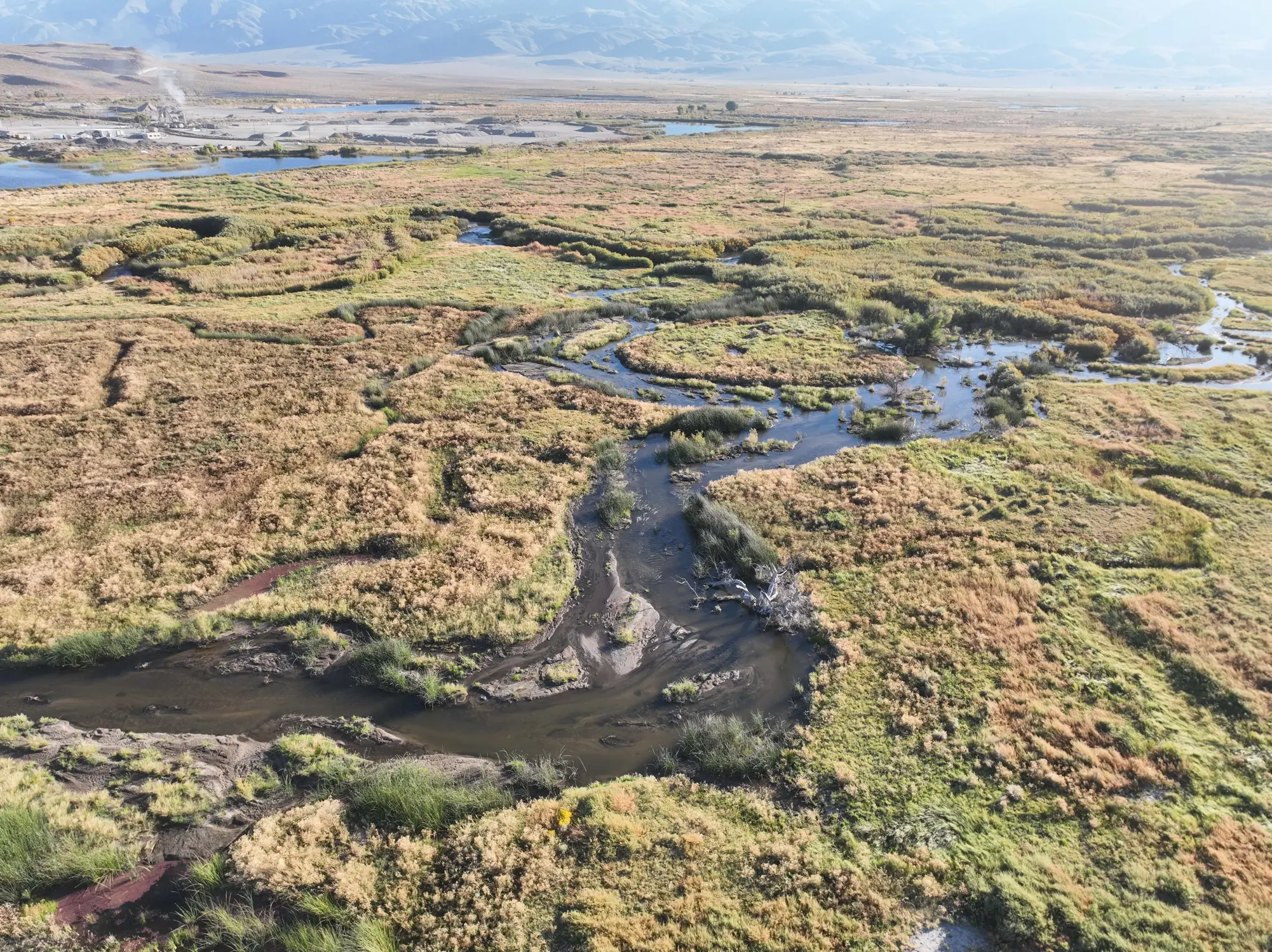 Five A winding stream flows through a dry, grassy landscape with distant mountains under a hazy sky.