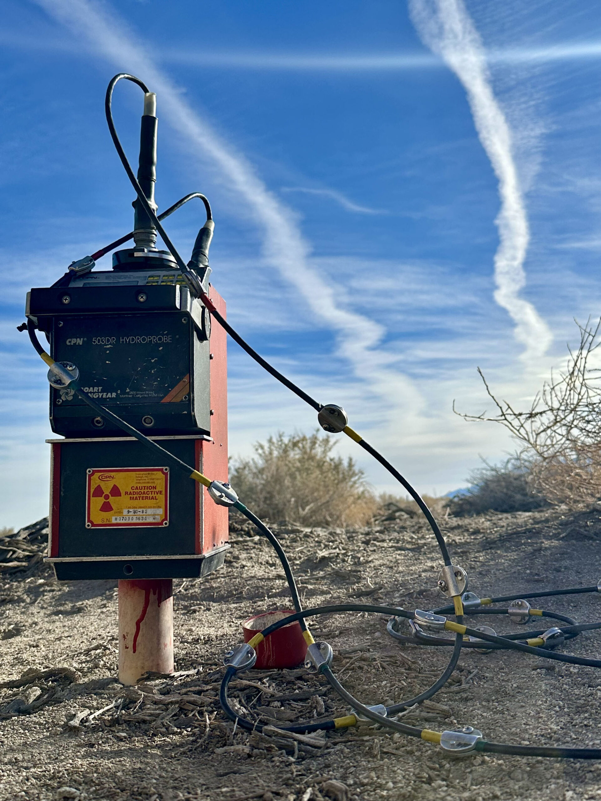 Neutron Probe Holly Alpert A red geophysical device with wires stands outdoors on dry soil under a blue sky with white clouds.
