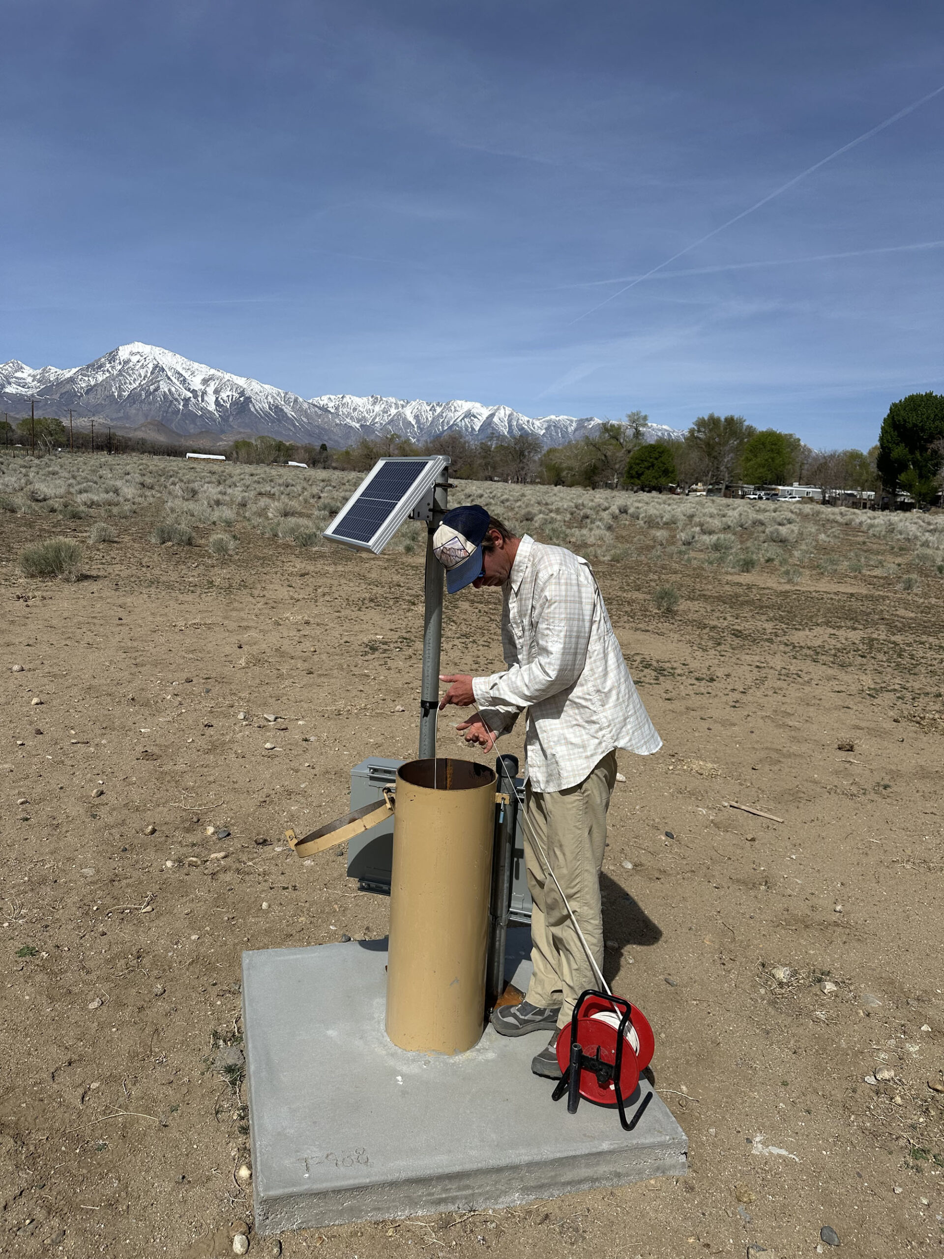 A person checks equipment at a water well site with mountains and a solar panel in the background.