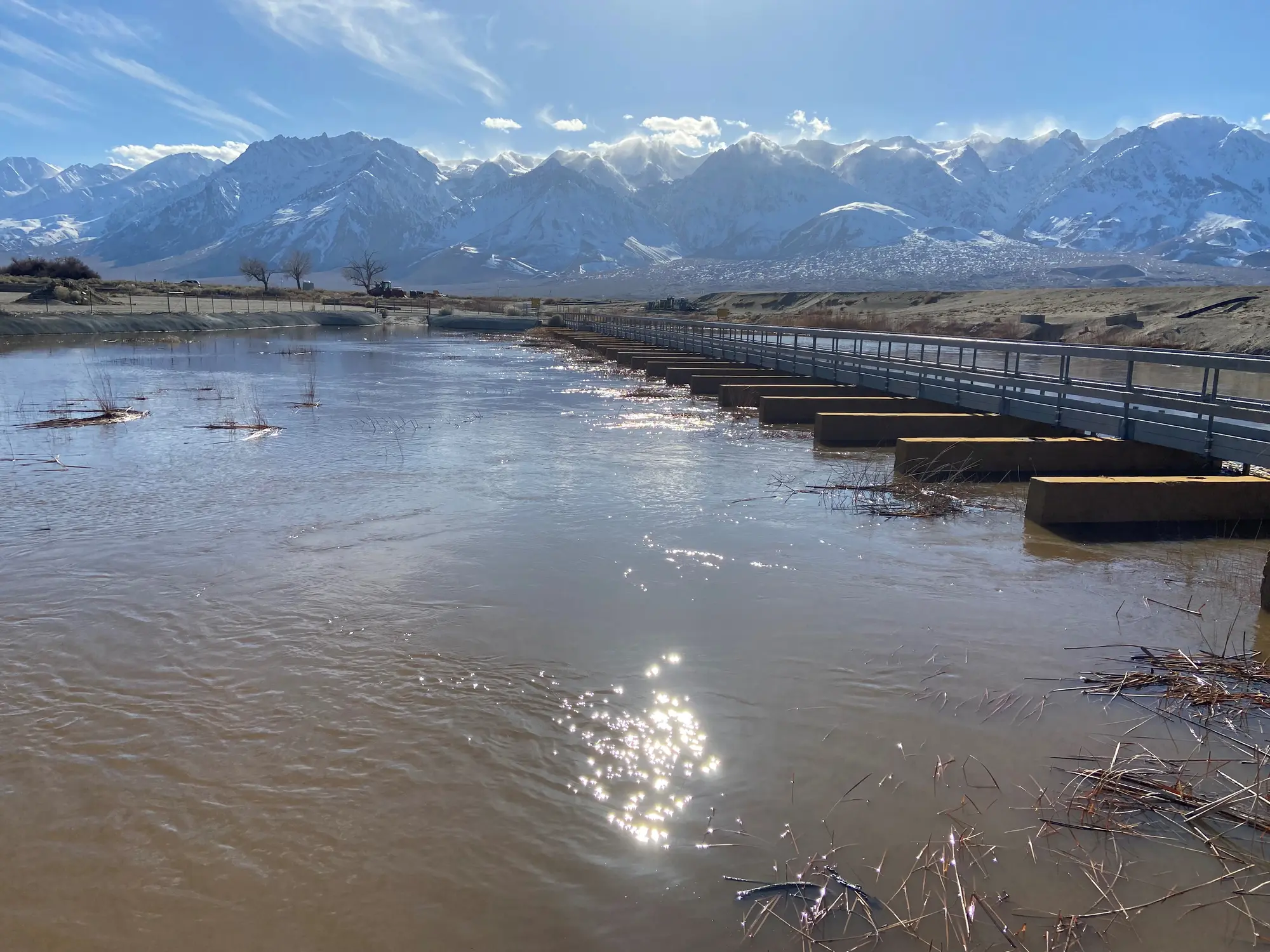 A flooded river reflects sunlight near a bridge, with snow-capped mountains in the background.